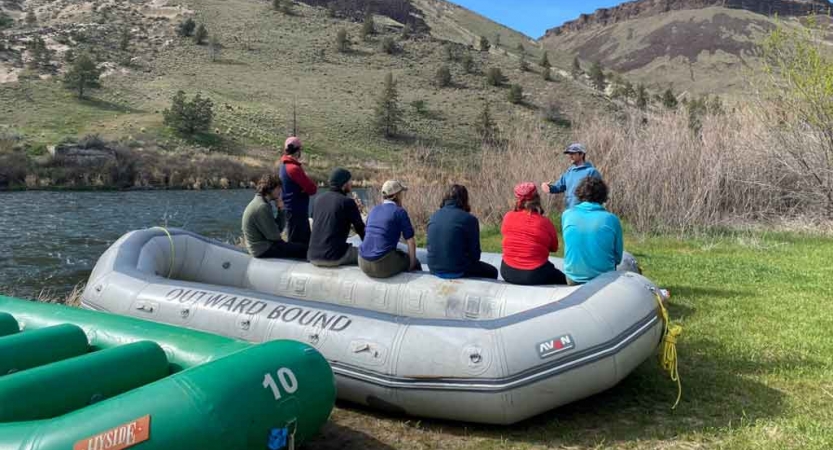 a group of gap year students rest on the side of a beached raft while listening to an instructor on an outward bound outdoor educator course